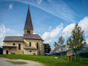 Eine kleine Steinkirche mit einem hohen Kirchturm steht auf einem grasbewachsenen Hügel unter einem blauen Himmel mit vereinzelten Wolken. In der Nähe befinden sich Bäume und ein hölzernes Hinweisschild, und im Hintergrund sind Berge zu sehen.