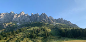 Zerklüftete Berggipfel erheben sich dramatisch über einen üppig grünen, mit Kiefern bestandenen Hang unter einem klaren blauen Himmel. Das Sonnenlicht beleuchtet die felsigen Gipfel und wirft Schatten in die Landschaft.