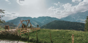 Eine rustikale Holzschaukel steht auf einer grasbewachsenen Hügelkuppe mit Blick auf üppig grüne Berge und Täler unter einem teilweise bewölkten Himmel. Ein Holzzaun führt am Rand entlang und umrahmt die malerische Landschaft.