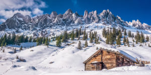 Eine rustikale Holzhütte steht an einem verschneiten, mit Kiefern bestandenen Hang, im Hintergrund erheben sich schroffe, felsige Berggipfel unter einem strahlend blauen Himmel mit vereinzelten Wolken.