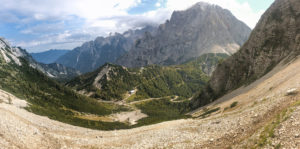 Ein Panoramablick auf eine kurvenreiche Straße durch ein üppig grünes Tal, umgeben von felsigen, hoch aufragenden Bergen unter einem teilweise bewölkten Himmel.